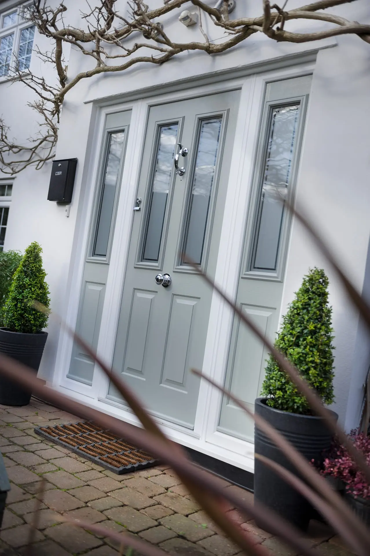 A pale grey composite front door with glazed side panels and chrome furniture, beside potted topiary