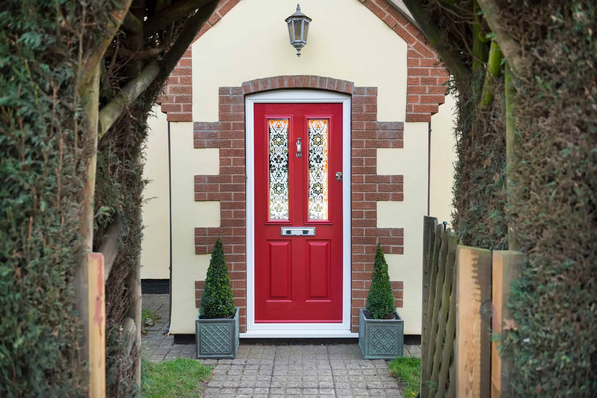 A red composite front door with decorative glazing between topiary trees